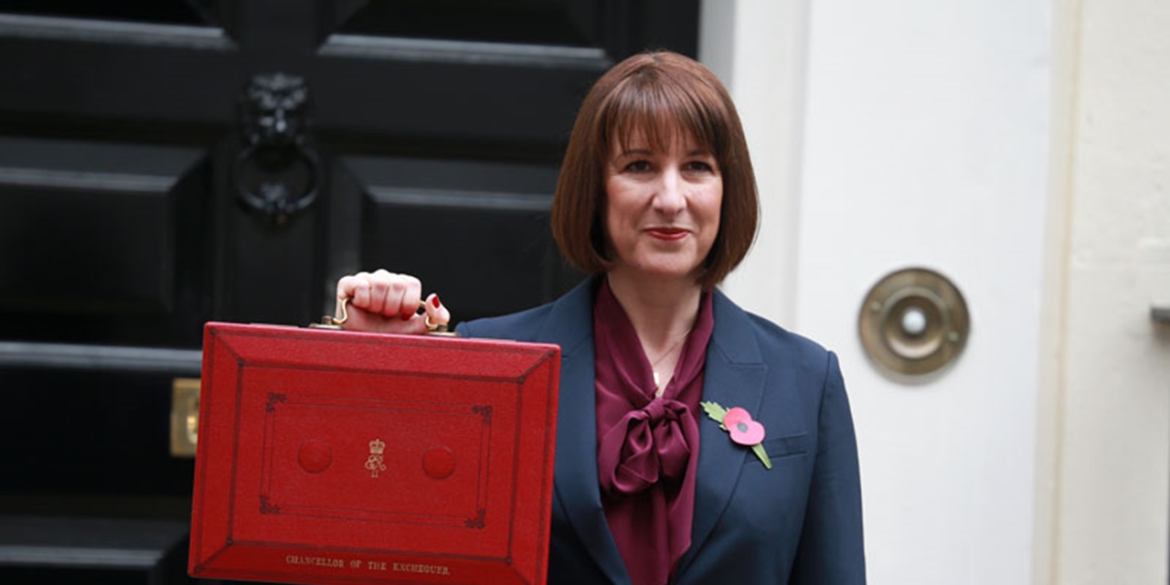 Chancellor of the Exchequer Rachel Reeves in a dark suit holding up a traditional red Budget box outside a black door, with a poppy pinned to her lapel