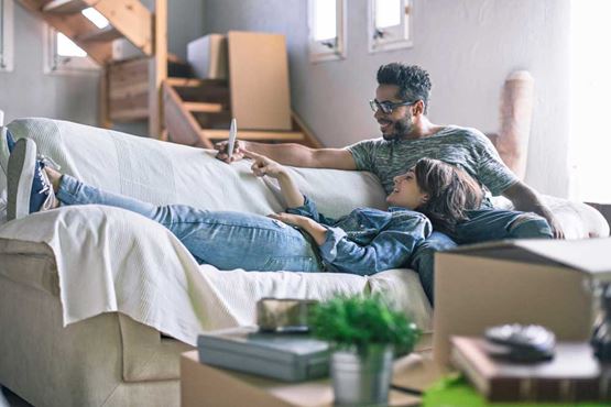 A young couple relax on a sofa while discussing something on a mobile phone screen - they are surrounded by boxes suggesting they may have just moved in
