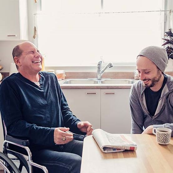 Photo of two men laughing around a kitchen table, one evidently a wheelchair user
