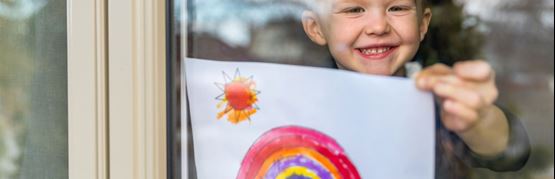 Child in window holding up a drawing of a rainbow