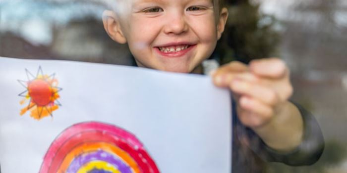 Child in window holding up a drawing of a rainbow