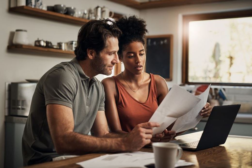 A couple at home sit at the kitche table looking at a laptop and various paper bills