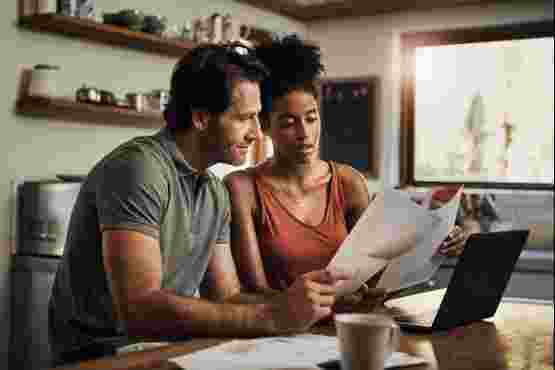 A couple at home sit at the kitche table looking at a laptop and various paper bills