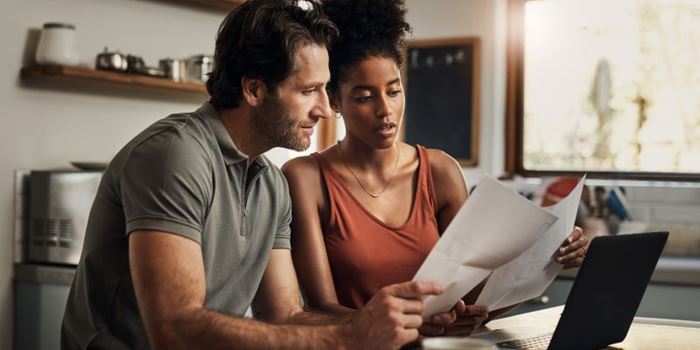 A couple at home sit at the kitche table looking at a laptop and various paper bills