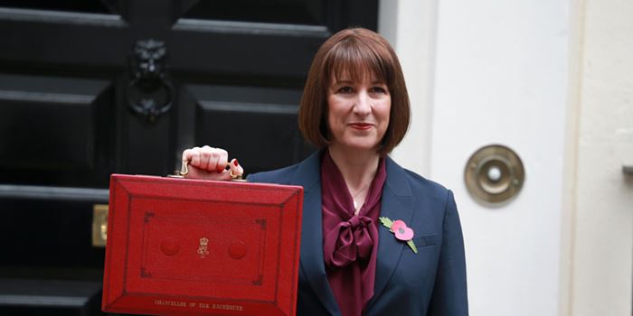 Chancellor of the Exchequer Rachel Reeves in a dark suit holding up a traditional red Budget box outside a black door, with a poppy pinned to her lapel