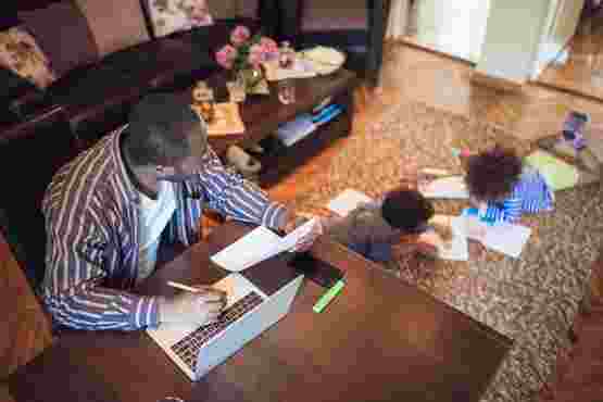 Man sat at desk at home with a laptop and a piece of paper while two children play on the rug behind him