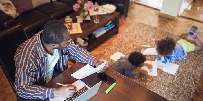 Man sat at desk at home with a laptop and a piece of paper while two children play on the rug behind him