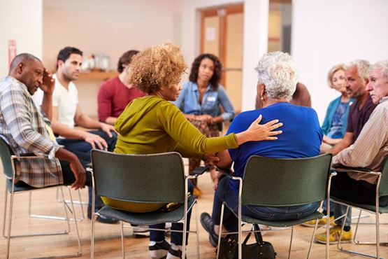 A group of people sit in a circle, focused on someone with her their back to the camera who is being consoled by her neighbour