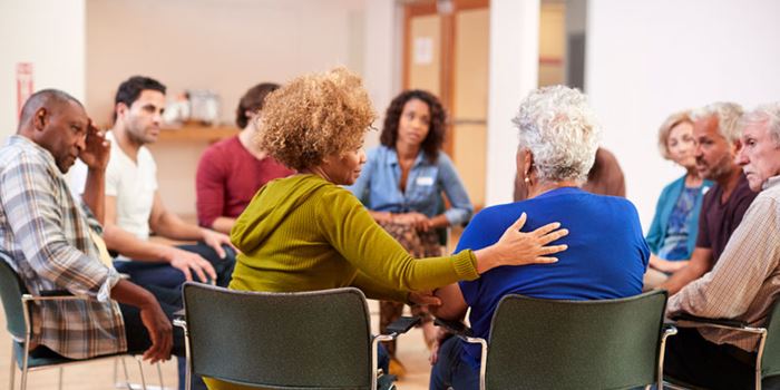 A group of people sit in a circle, focused on someone with her their back to the camera who is being consoled by her neighbour