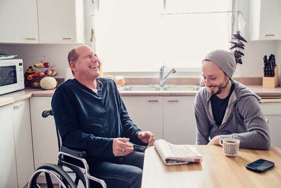 Two men in a kitchen, sat at the table with a coffee and a newspaper, laughing