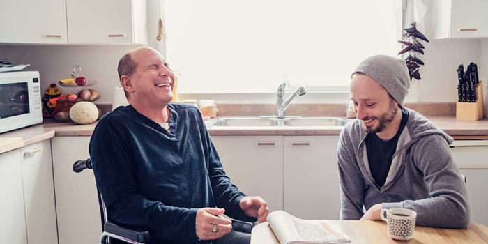 Two men in a kitchen, sat at the table with a coffee and a newspaper, laughing