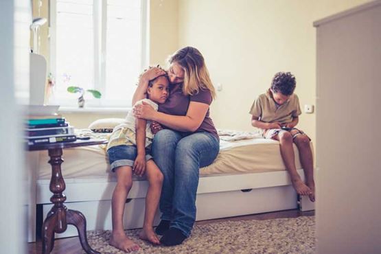 A woman sits on a bed with two children, holding one close and looking very concerned