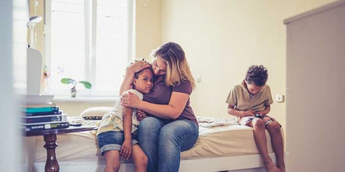 A woman sits on a bed with two children, holding one close and looking very concerned