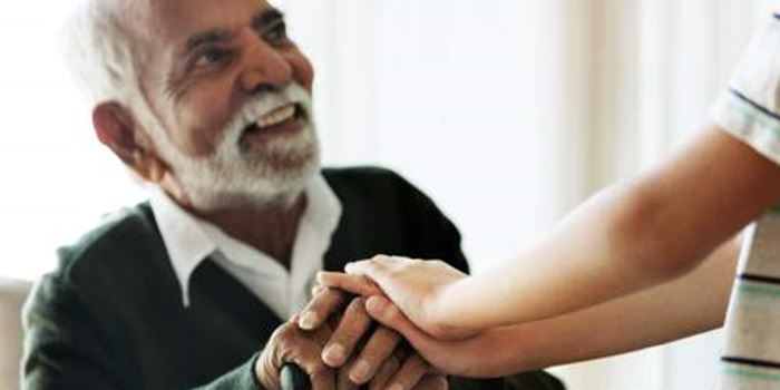 An older man sits indoors smiling up at someone. His hands rest on his walking stick. Their hands rest on his.