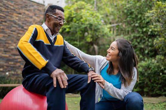 In a garden, a smiling man sits on an exercise ball supported by a woman crouched down in front of him