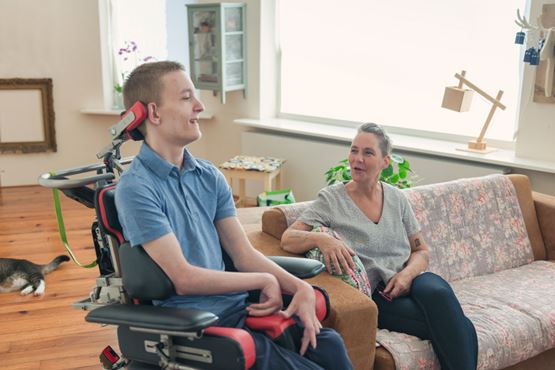 A young man in a wheel chair smiles while a woman sat next to him speaks and smiles
