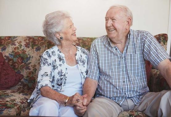 An older couple sit close on a sofa holding hands and smiling at one another