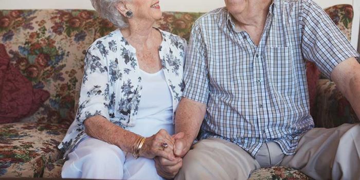 An older couple sit close on a sofa holding hands and smiling at one another