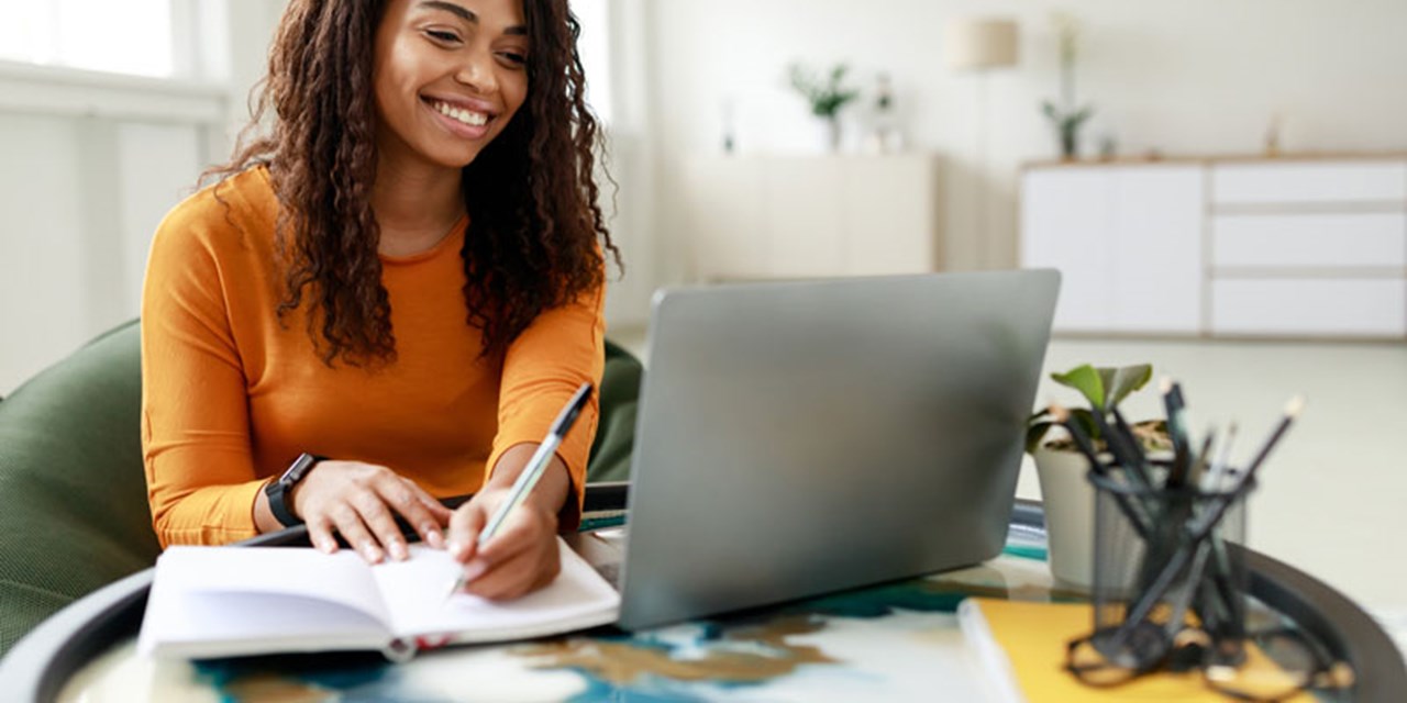 Person sat at table smiling at a laptop and taking notes