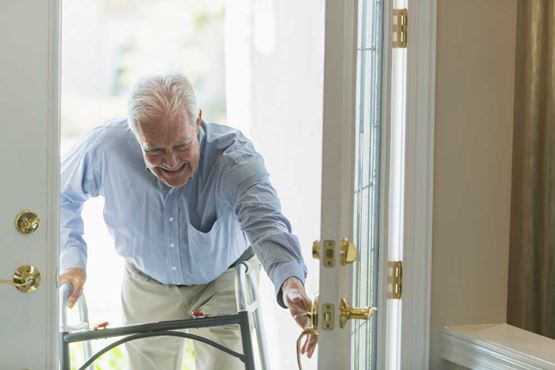 An older man with a walking frame smiles as he arrives home through the front door