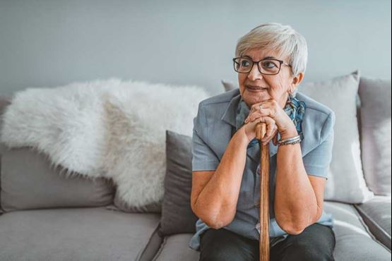 An older woman sat on sofa resting her hands and chin on the top of a walking stick