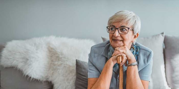 An older woman sat on sofa resting her hands and chin on the top of a walking stick